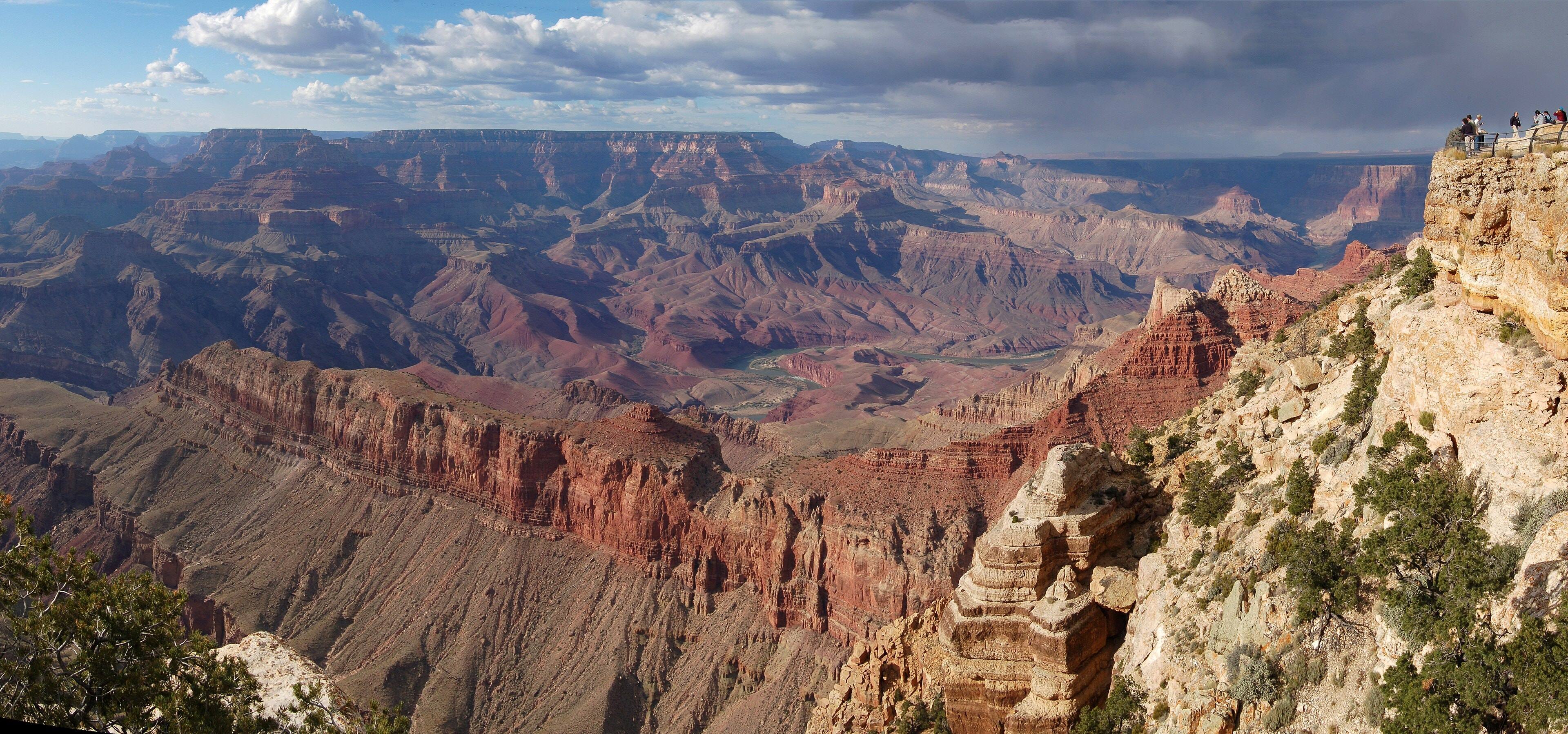 Layered red and tan canyon walls seen from Lipan Point, with a bend of the Colorado River far below under a cloudy sky